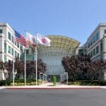 Apple Headquarters in Cupertino, A modern office building with a glass entrance, light-colored facade, and symmetrical design is shown. Three flags—the American flag, a white flag, and a California state flag—are flying in front. Trees and bushes line the walkway leading to the entrance.