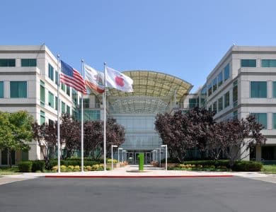 Apple Headquarters in Cupertino, A modern office building with a glass entrance, light-colored facade, and symmetrical design is shown. Three flags—the American flag, a white flag, and a California state flag—are flying in front. Trees and bushes line the walkway leading to the entrance.
