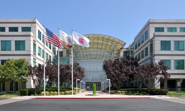 Apple Headquarters in Cupertino, A modern office building with a glass entrance, light-colored facade, and symmetrical design is shown. Three flags—the American flag, a white flag, and a California state flag—are flying in front. Trees and bushes line the walkway leading to the entrance.