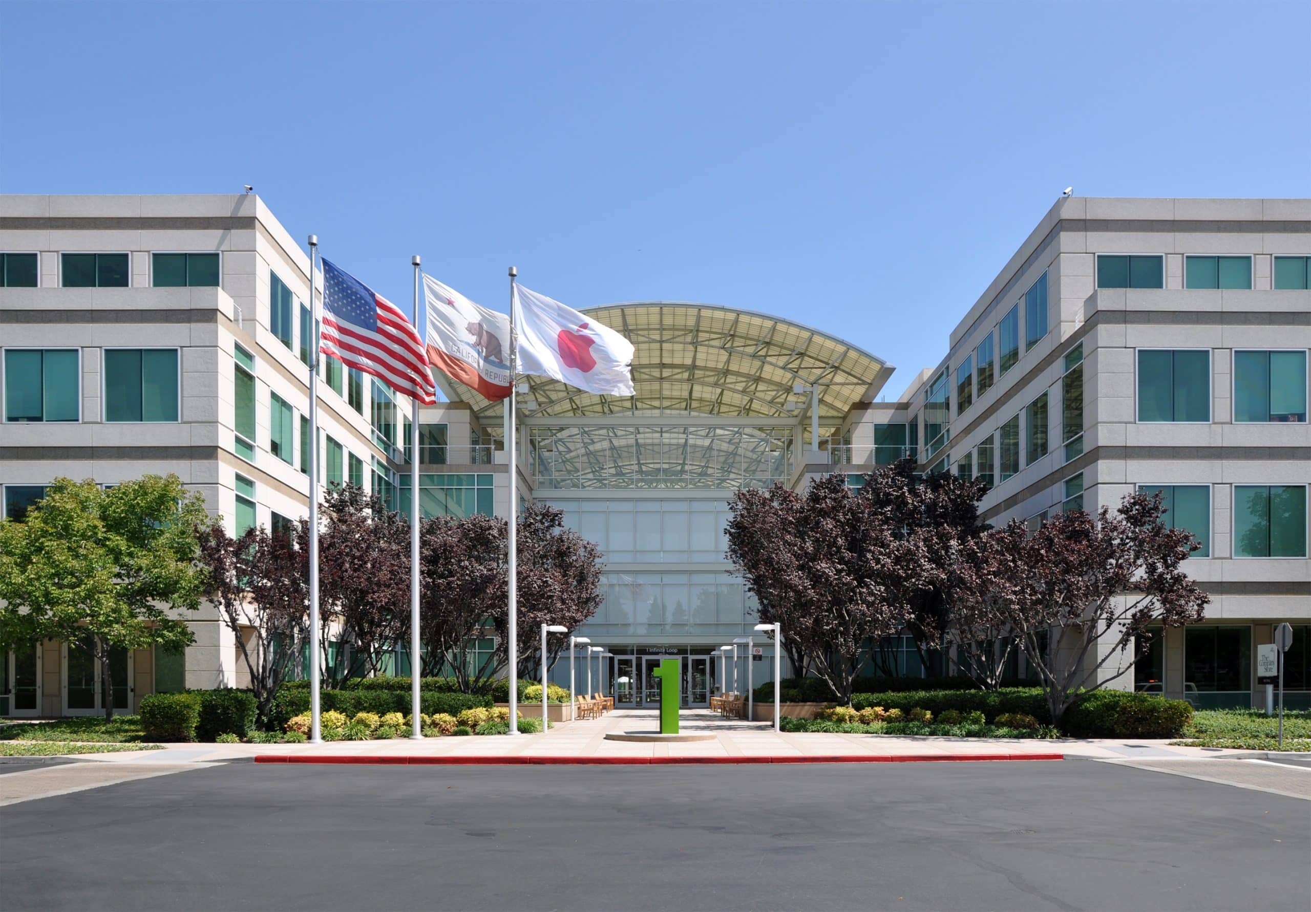 Apple Headquarters in Cupertino, A modern office building with a glass entrance, light-colored facade, and symmetrical design is shown. Three flags—the American flag, a white flag, and a California state flag—are flying in front. Trees and bushes line the walkway leading to the entrance.