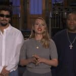 Three people stand indoors. The man on the left, reminiscent of Bad Bunny SNL, has curly hair, a beard, sunglasses, and a white shirt. The woman in the center wears red lipstick and jewelry. The man on the right wears a dark shirt and bold cross necklace.