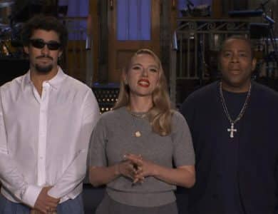 Three people stand indoors. The man on the left, reminiscent of Bad Bunny SNL, has curly hair, a beard, sunglasses, and a white shirt. The woman in the center wears red lipstick and jewelry. The man on the right wears a dark shirt and bold cross necklace.