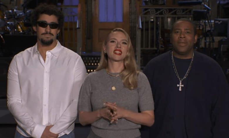 Three people stand indoors. The man on the left, reminiscent of Bad Bunny SNL, has curly hair, a beard, sunglasses, and a white shirt. The woman in the center wears red lipstick and jewelry. The man on the right wears a dark shirt and bold cross necklace.