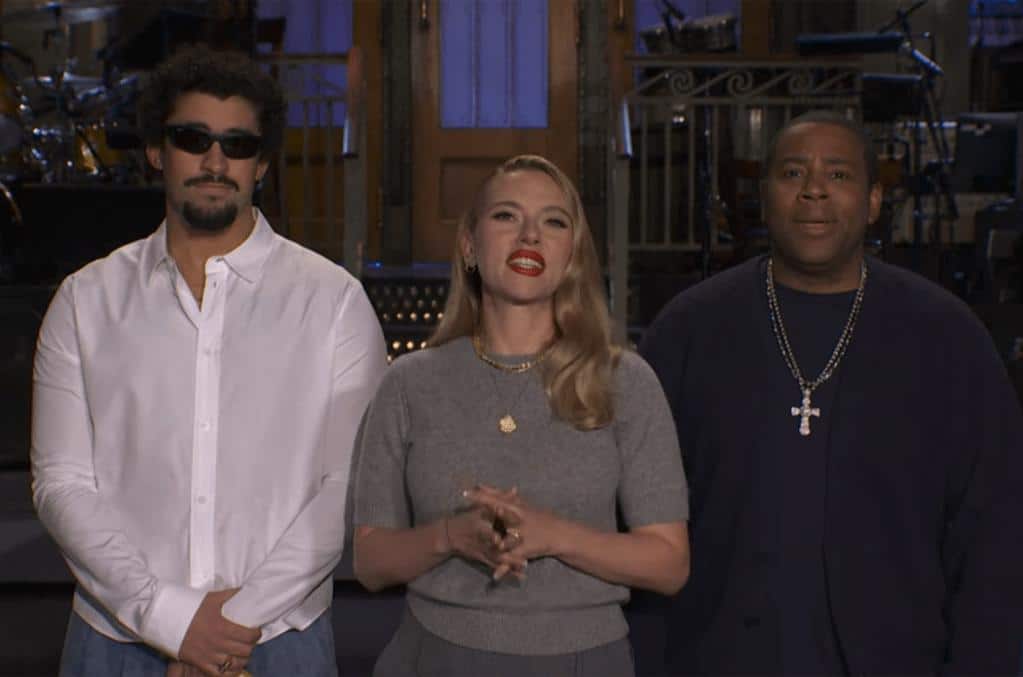 Three people stand indoors. The man on the left, reminiscent of Bad Bunny SNL, has curly hair, a beard, sunglasses, and a white shirt. The woman in the center wears red lipstick and jewelry. The man on the right wears a dark shirt and bold cross necklace.