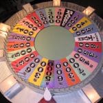 A colorful, top-down view of the iconic Wheel of Fortune game wheel, featuring various cash values, “BANKRUPT,” “LOSE A TURN,” and prize wedges. The wheel is divided into many colored segments with white flaps at regular intervals around the edge.