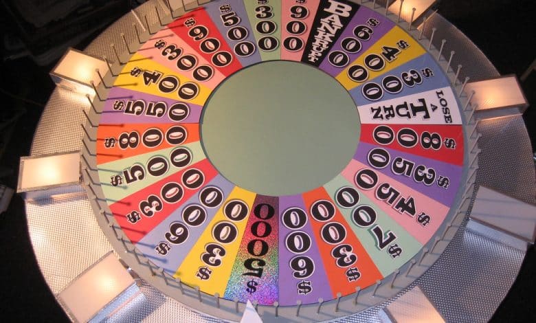 A colorful, top-down view of the iconic Wheel of Fortune game wheel, featuring various cash values, “BANKRUPT,” “LOSE A TURN,” and prize wedges. The wheel is divided into many colored segments with white flaps at regular intervals around the edge.