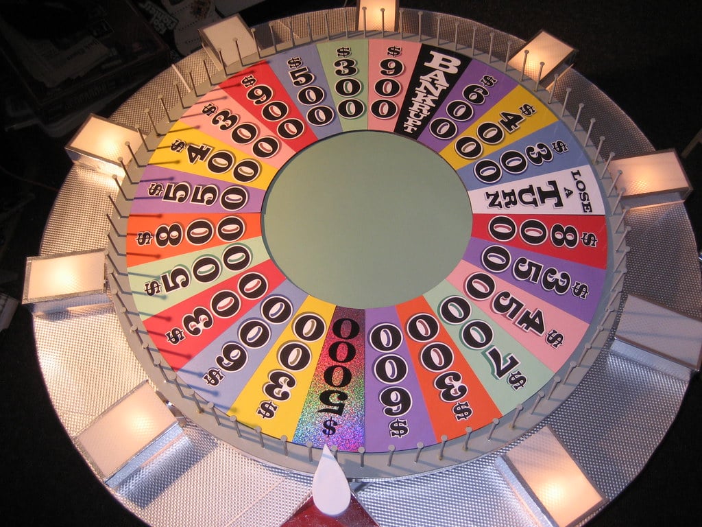 A colorful, top-down view of the iconic Wheel of Fortune game wheel, featuring various cash values, “BANKRUPT,” “LOSE A TURN,” and prize wedges. The wheel is divided into many colored segments with white flaps at regular intervals around the edge.