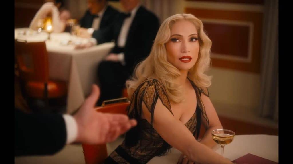 A glamorous woman with wavy blonde hair and bold red lipstick sits at a restaurant table in an elegant black and gold dress, evoking Kiss of the Spider Woman, as she holds a cocktail and looks up at a tuxedoed man extending his hand.