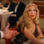 A glamorous woman with wavy blonde hair and bold red lipstick sits at a restaurant table in an elegant black and gold dress, evoking Kiss of the Spider Woman, as she holds a cocktail and looks up at a tuxedoed man extending his hand.