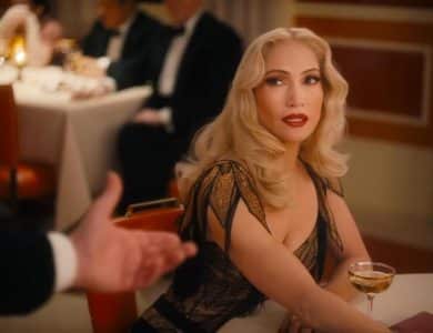 A glamorous woman with wavy blonde hair and bold red lipstick sits at a restaurant table in an elegant black and gold dress, evoking Kiss of the Spider Woman, as she holds a cocktail and looks up at a tuxedoed man extending his hand.
