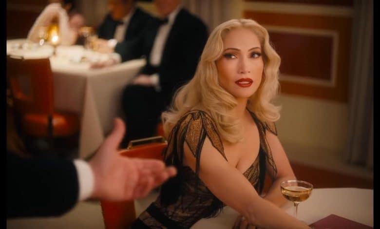 A glamorous woman with wavy blonde hair and bold red lipstick sits at a restaurant table in an elegant black and gold dress, evoking Kiss of the Spider Woman, as she holds a cocktail and looks up at a tuxedoed man extending his hand.