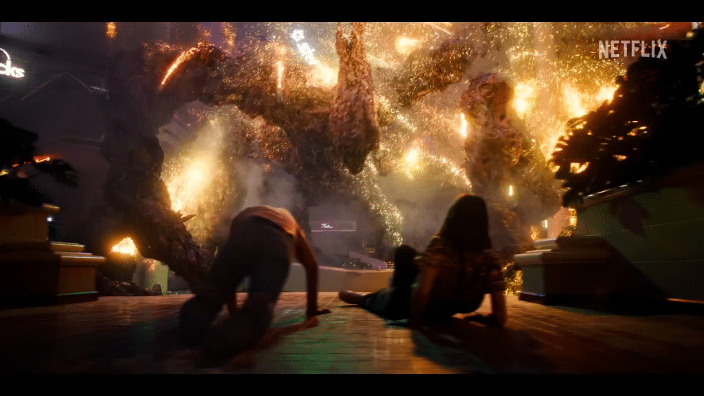 Two people crawl away on the floor as a massive, glowing, monstrous creature with outstretched arms and fiery, golden sparks looms over them inside a building—a dramatic scene straight from Stranger Things. The Netflix logo appears in the upper right corner.
