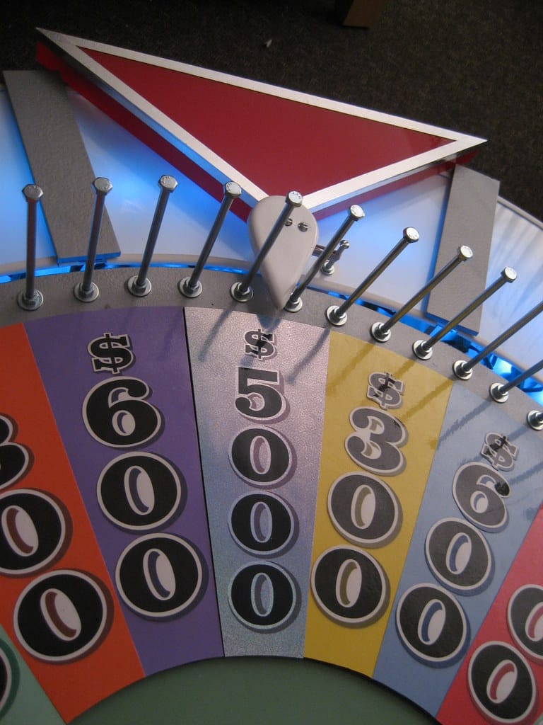 Close-up of a Wheel of Fortune-style game wheel displaying numbered dollar amounts ($6,000, $5,000, $3,000) in colored wedges. A large arrow points downward as metal pegs separate each vibrant segment.